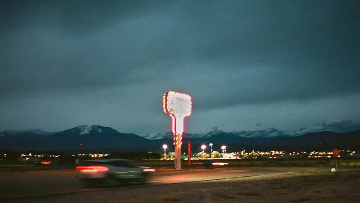 Neon-lit roadside sign glowing against dark mountains at dusk with car light trails.