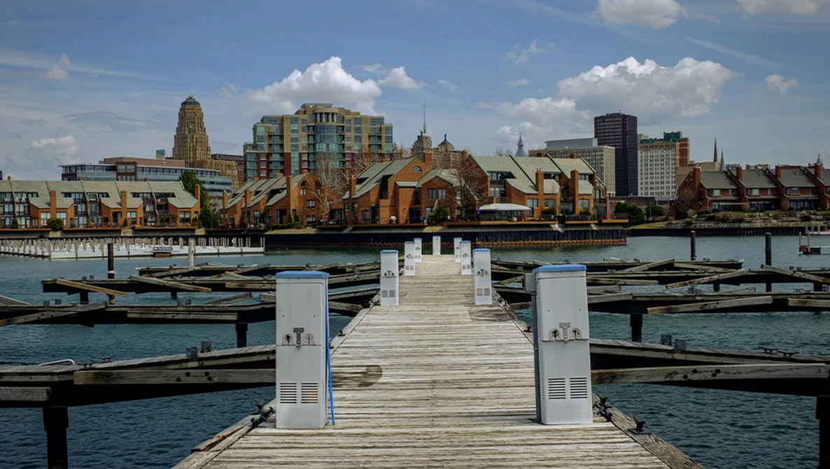 Wooden pier extending into turquoise water with city skyline featuring historic and modern buildings in background.
