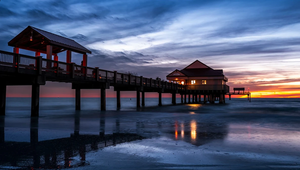 Illuminated pier structures silhouetted against a vibrant sunset sky reflecting in shallow water.