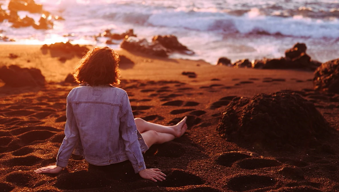 Person sitting on beach sand facing ocean at golden hour sunset.