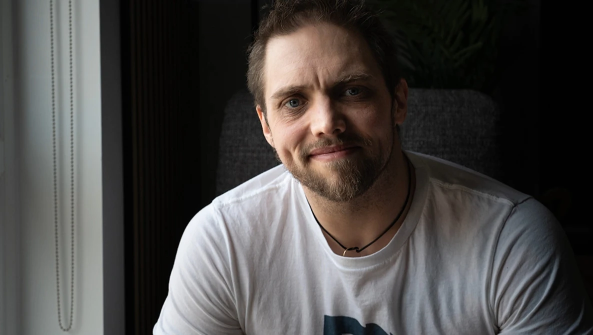 Man with goatee wearing light gray t-shirt, photographed against dark background with window light.