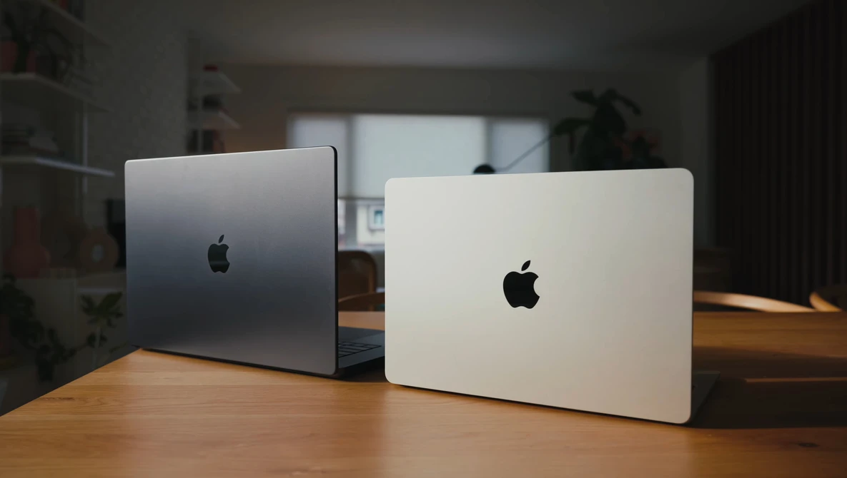 Two MacBook laptops displayed closed on a wooden desk, one silver and one white, photographed in indoor studio lighting.