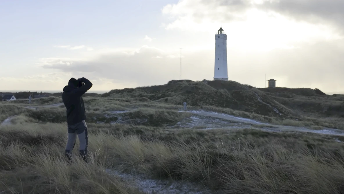 Photographer with camera raised to face standing in coastal dunes with lighthouse on distant hill.