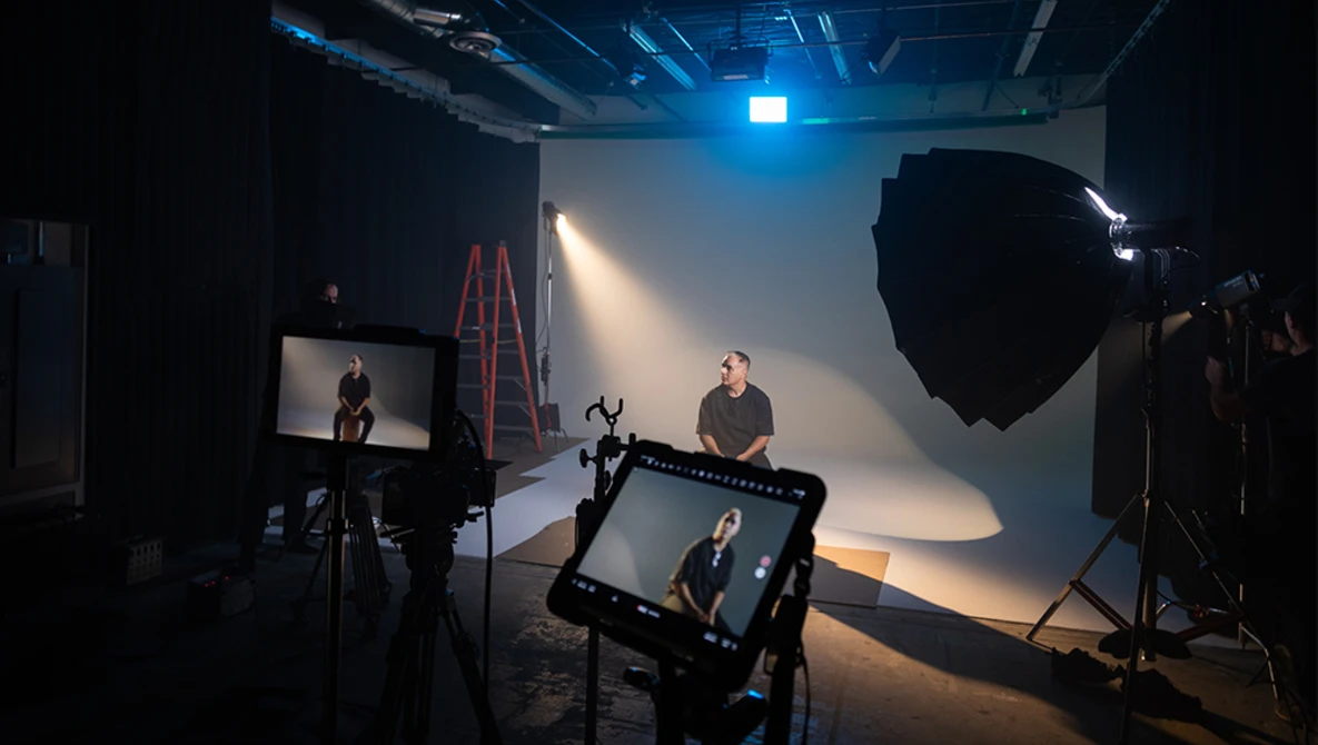 Professional film production studio setup with subject seated in key light, multiple cameras and monitors monitoring the shoot.