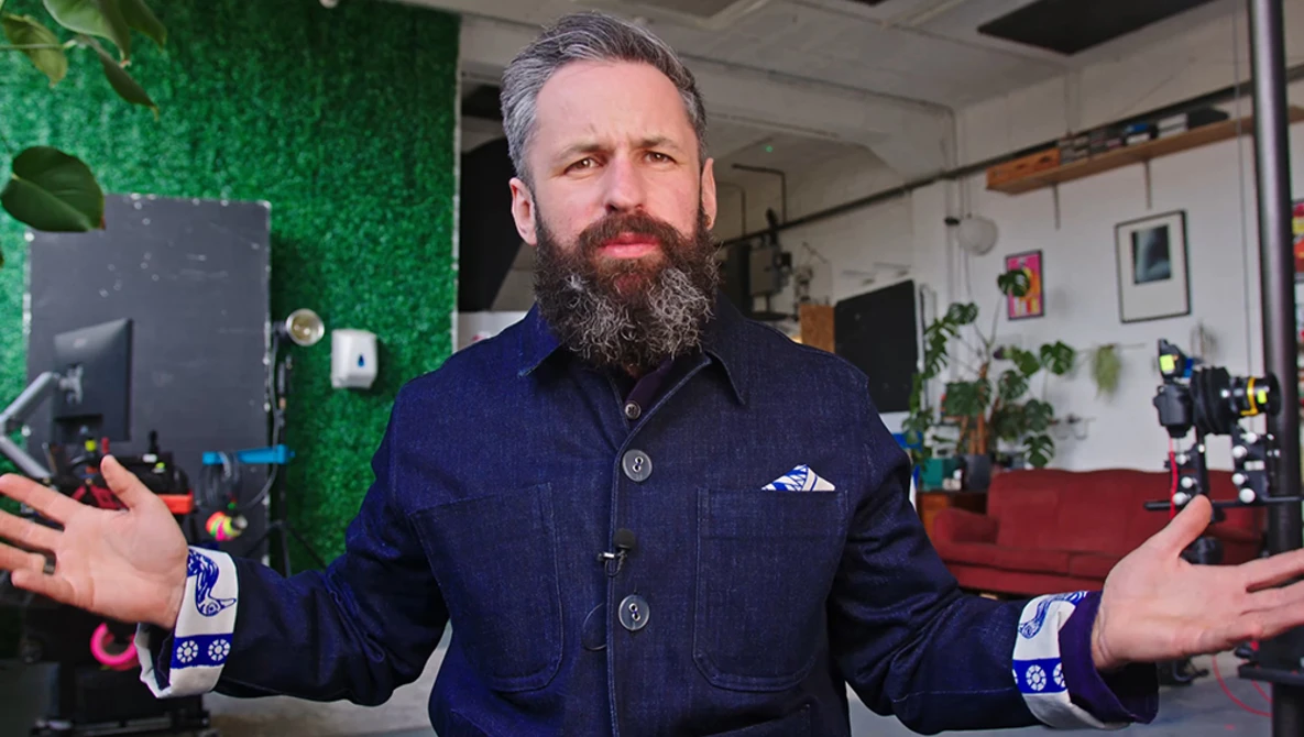 Bearded man in dark denim jacket gesturing expressively in a creative studio space.