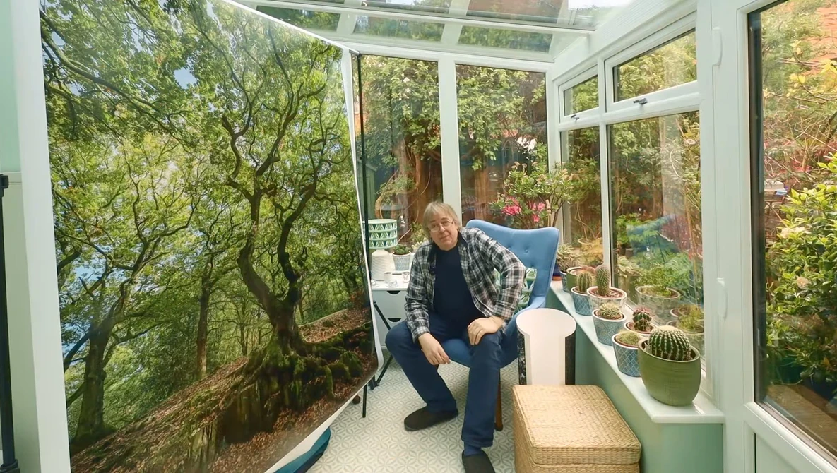 Man in plaid shirt crouching in sunlit conservatory with forest mural and potted plants.