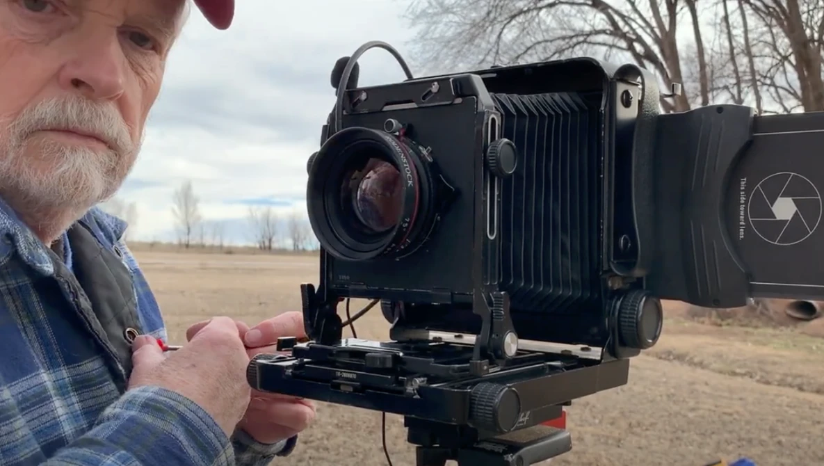Photographer holding a large format camera with bellows and ground glass in an open field.
