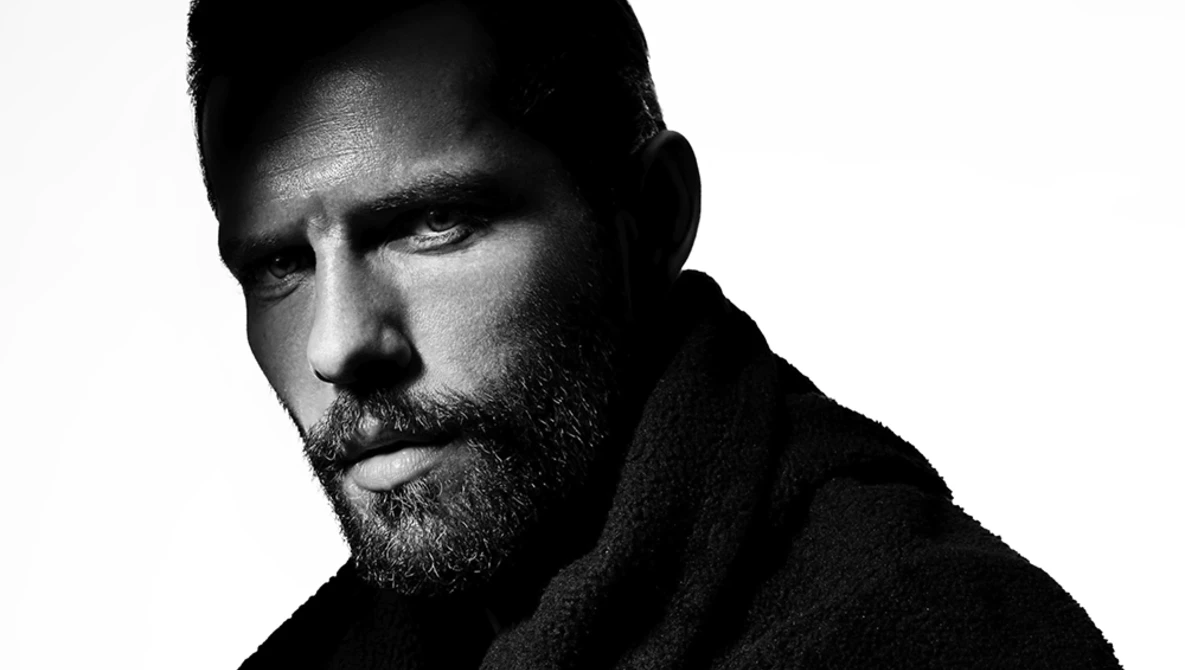 Bearded man with dark hair in black and white portrait photography, shot against white background with dramatic side lighting.