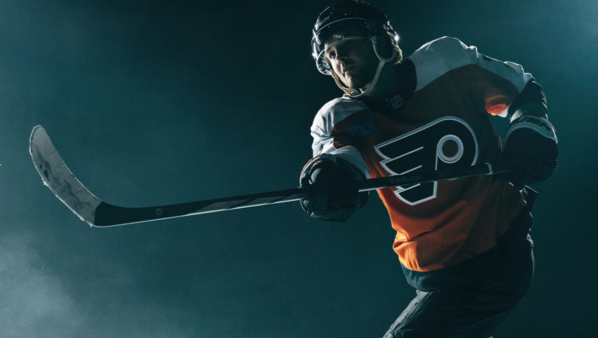 Hockey player in orange and black jersey holding stick against dark blue background with atmospheric lighting.