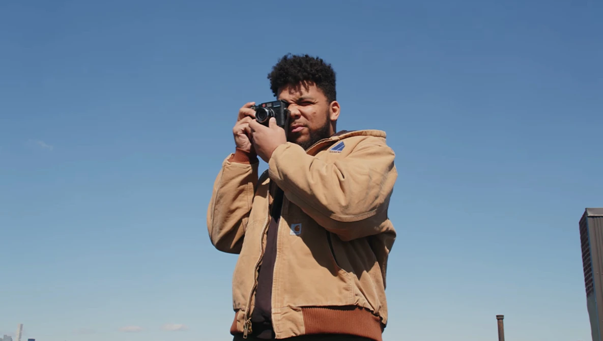 Photographer holding a camera up to his face while looking through the viewfinder against a clear blue sky.
