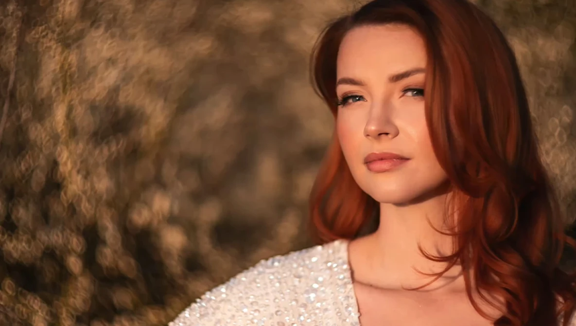 Woman with reddish-brown hair against a textured brown background, wearing a light-colored knit sweater.