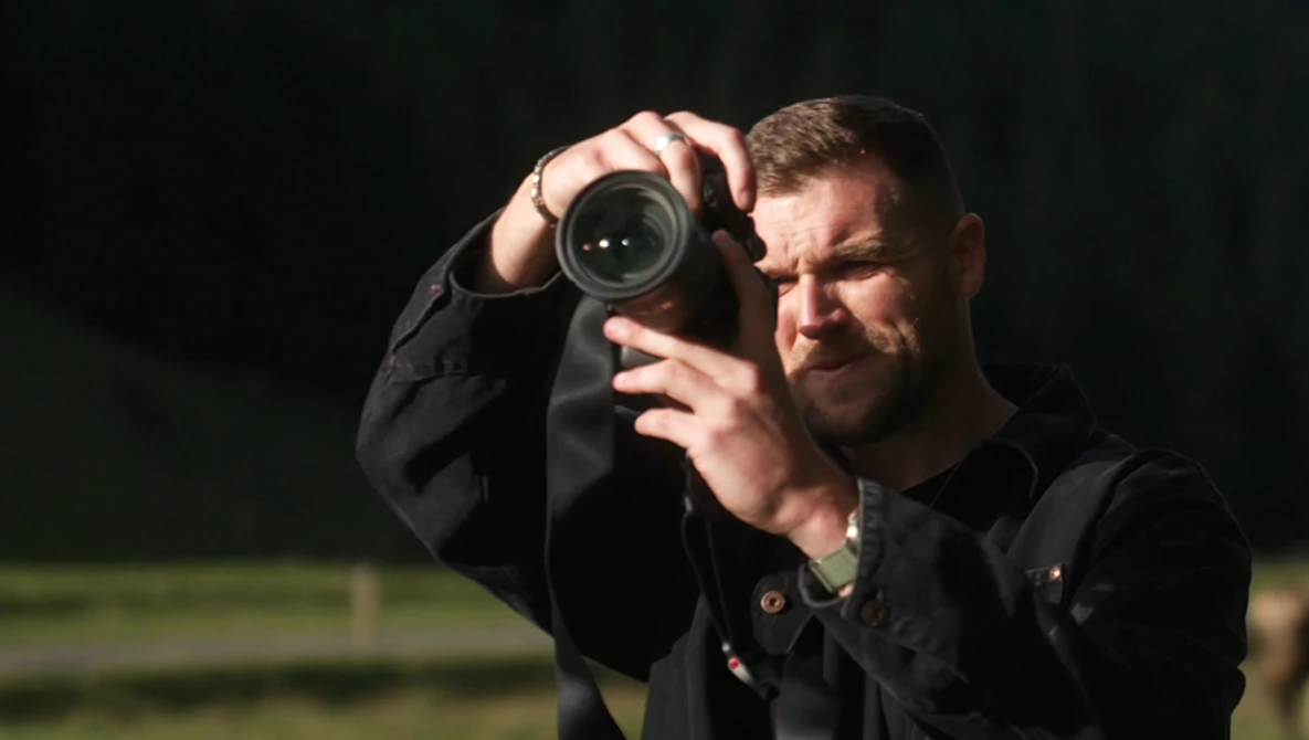 Photographer in black jacket holding camera with telephoto lens to his eye, focused on subject with concentrated expression.
