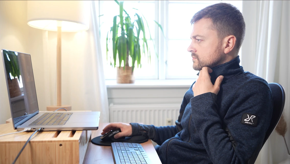 Man at desk reviewing laptop screen while working with keyboard and mouse in bright home office.