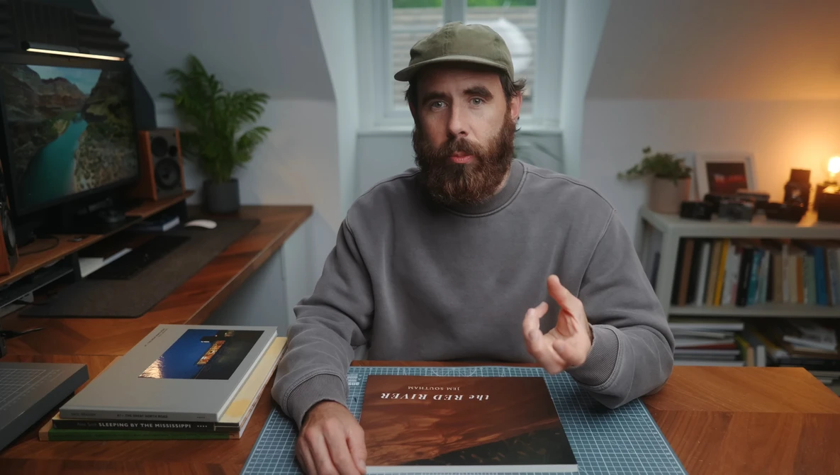 Bearded man wearing a gray sweatshirt and cap gestures while discussing photography books at a wooden desk.