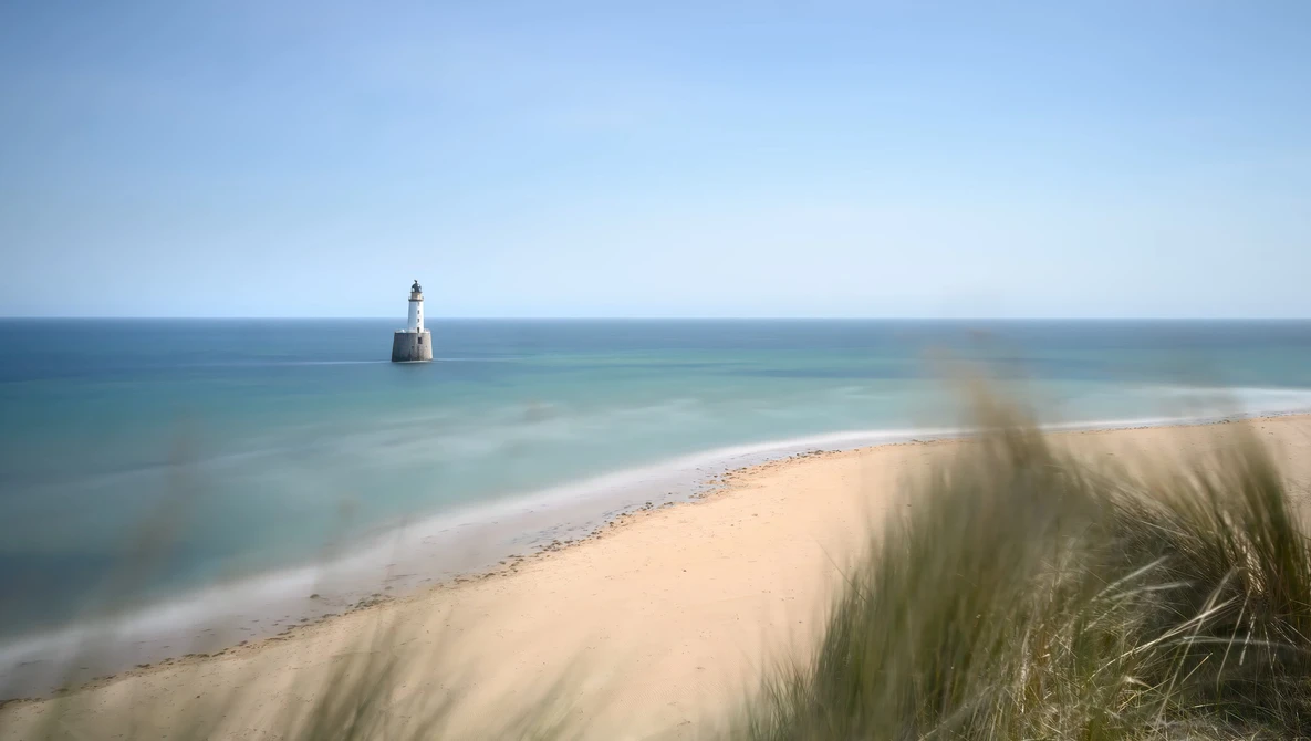 Sandy beach with dune grasses in foreground, calm turquoise water, and distant lighthouse on the horizon.