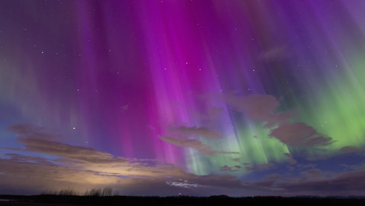 Aurora borealis display with vibrant magenta and green bands arcing across a twilight sky above a dark landscape.