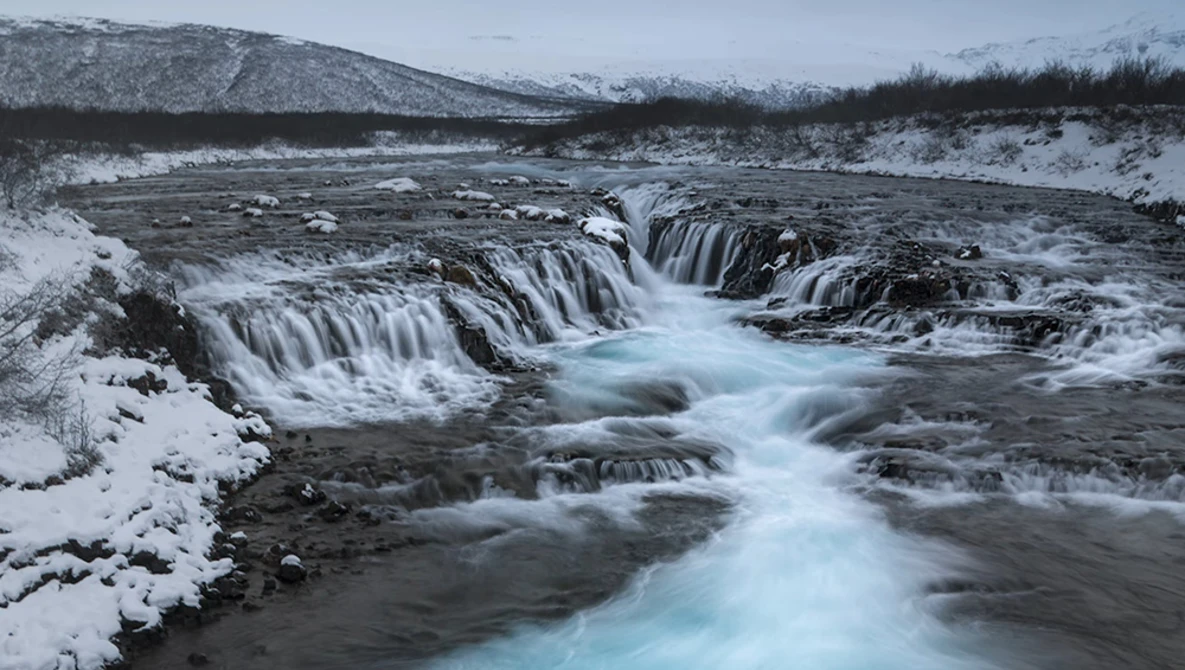 Frozen waterfall cascading through snow-covered landscape with turquoise glacial water flowing between dark rocks.