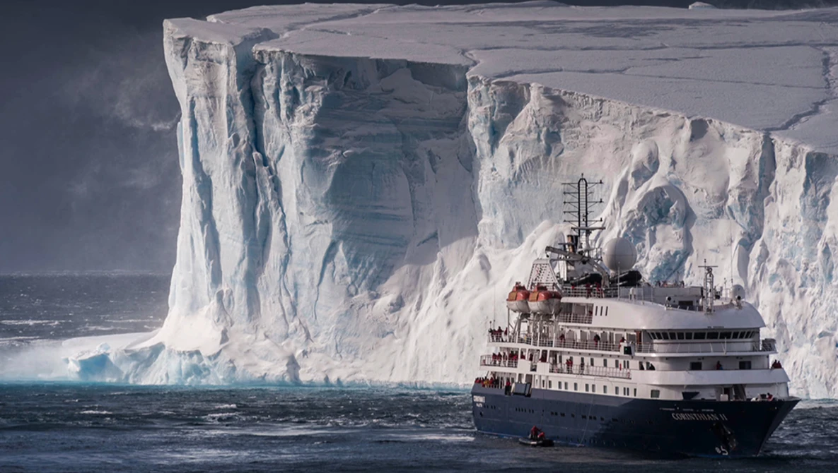 Research vessel positioned before a massive ice shelf with towering white cliffs and deep crevasses.