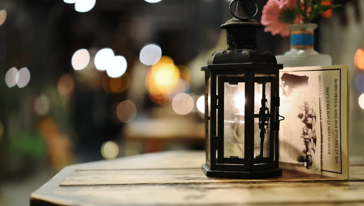 Black metal lantern with candle next to a book on wooden surface, with blurred bokeh lights in background.