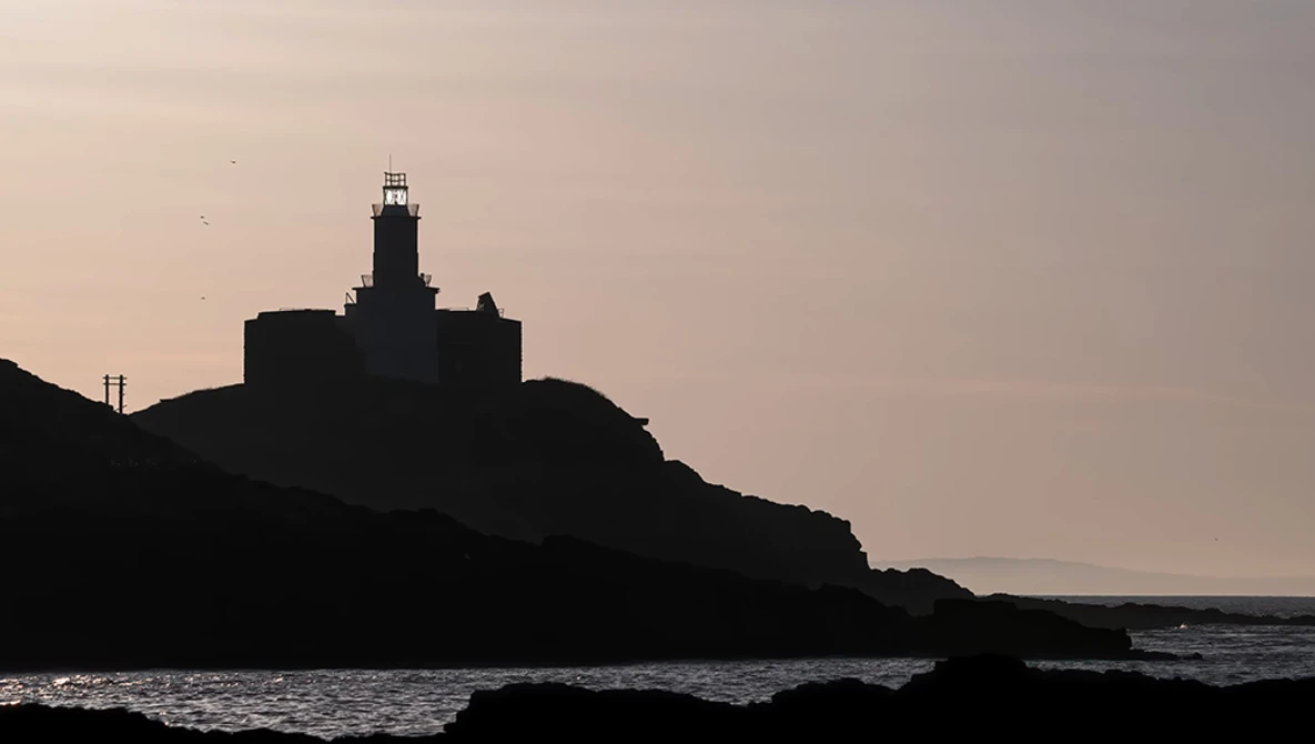 Lighthouse silhouetted against a twilight sky on a rocky coastal promontory.