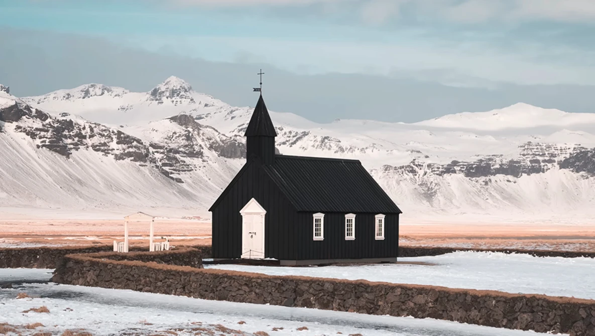 Black wooden church with white door and cross standing alone in snow-covered landscape beneath snow-capped mountains.