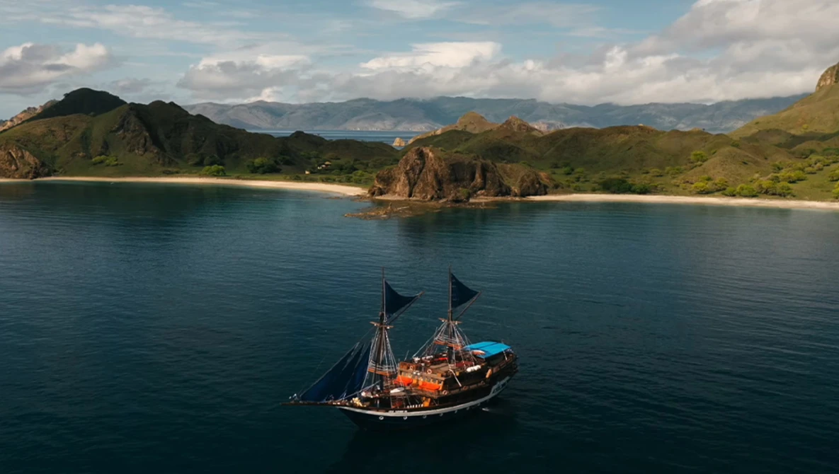 Traditional sailing vessel with dark hull and two masts anchored in calm turquoise water near a sandy beach and forested coastline.