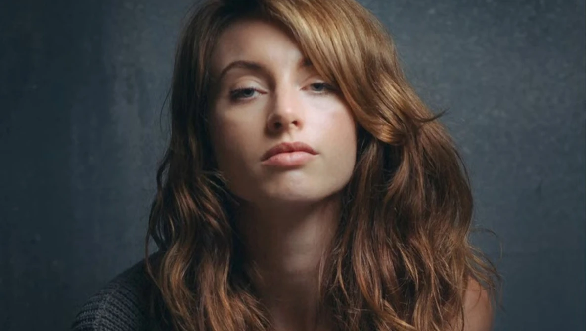 Woman with long wavy brown hair against a dark textured backdrop, looking downward with neutral expression.