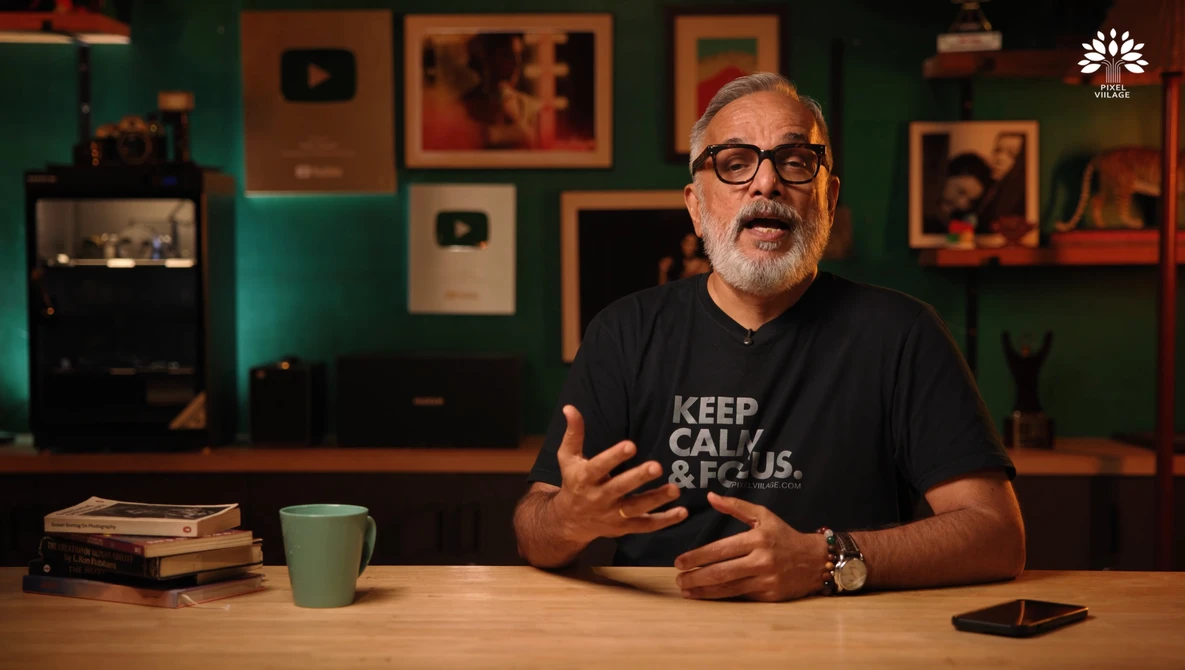 Man wearing glasses and black t-shirt gestures while speaking at wooden desk in studio setting.