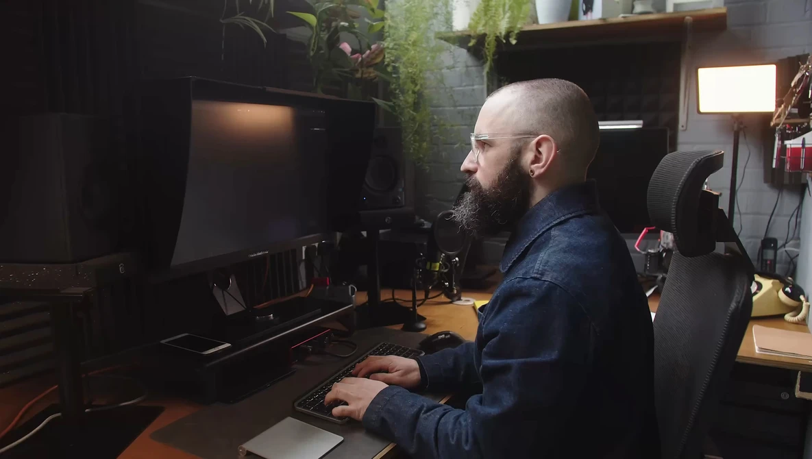 Bearded man working at a computer desk in a dark studio space surrounded by plants and equipment.