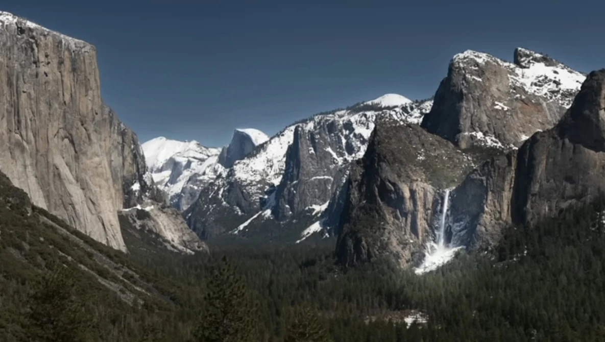 Snow-capped granite peaks and forested valley under clear blue sky with waterfalls.