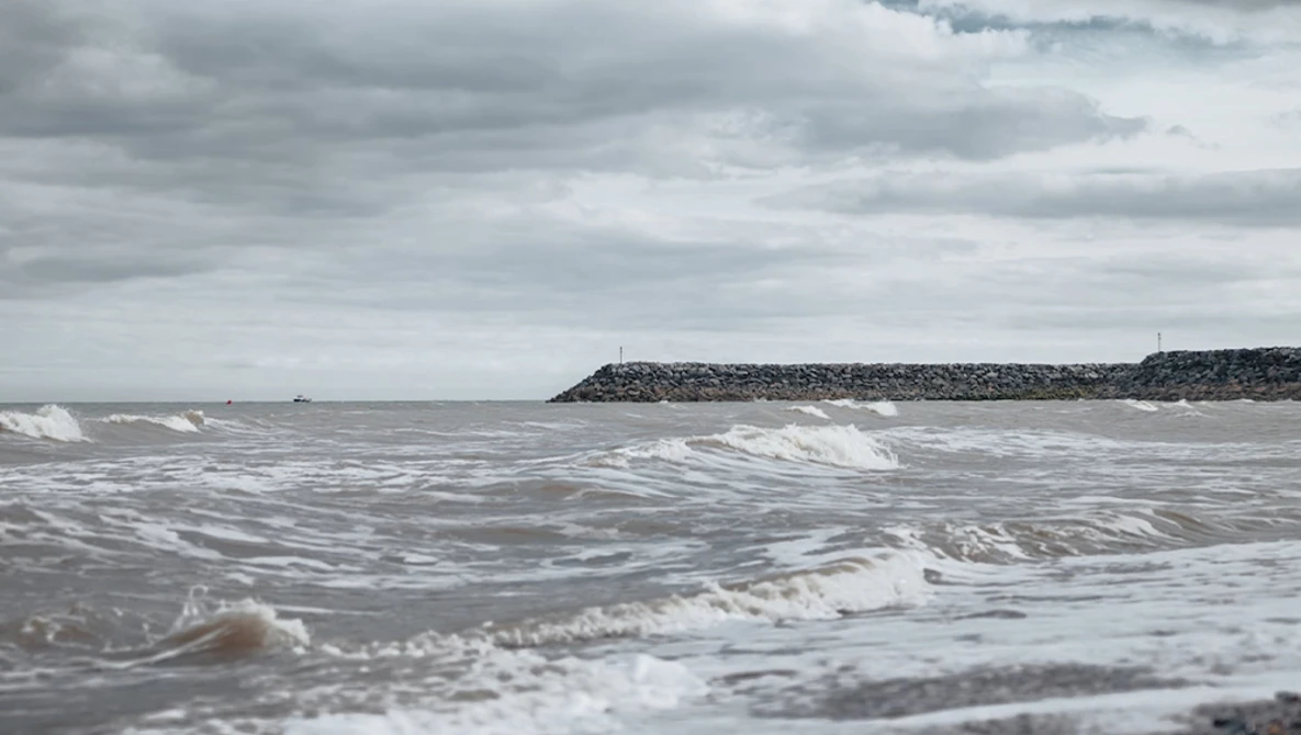 Rough ocean waves breaking on a beach under overcast sky with a rocky breakwater visible in the distance.