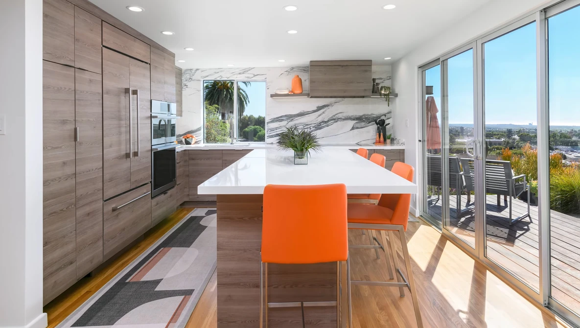 Modern kitchen with white marble countertops, wood cabinetry, and orange bar stools overlooking a city landscape through floor-to-ceiling windows.