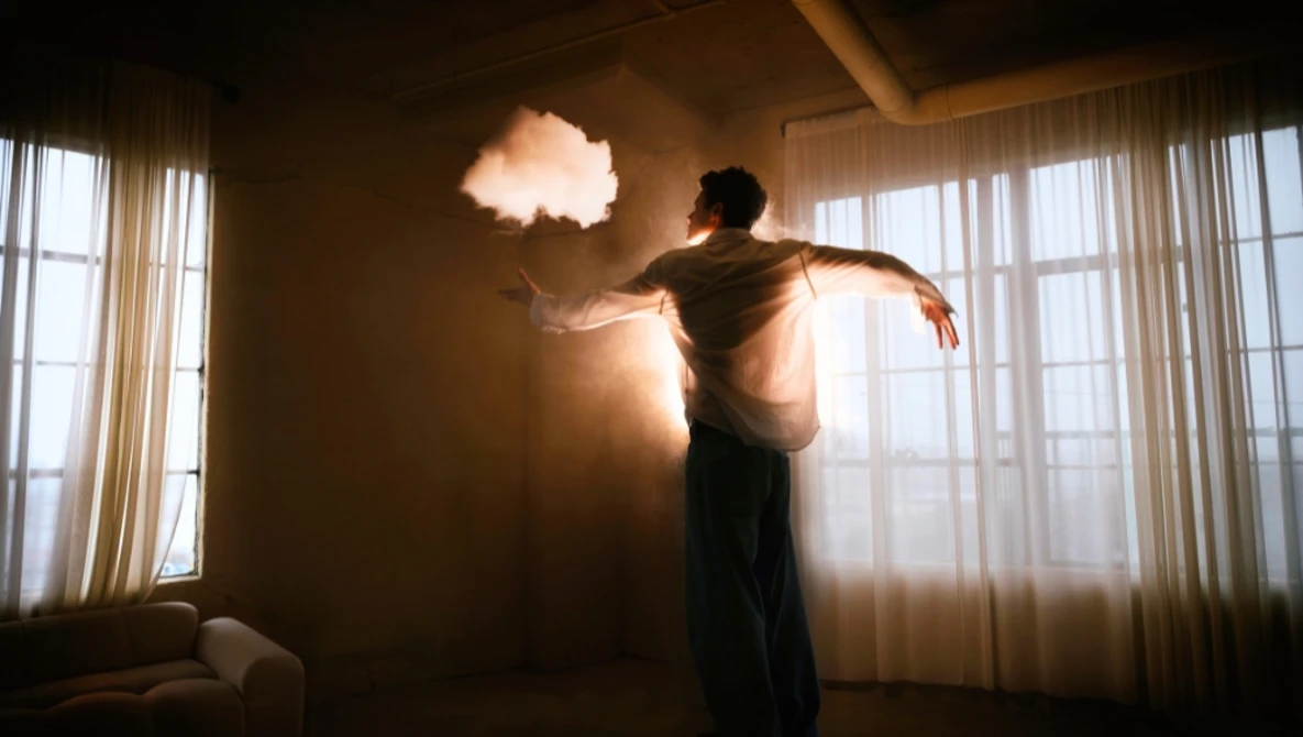 Person silhouetted against glowing cloud-shaped light suspended in a minimalist interior with large windows.