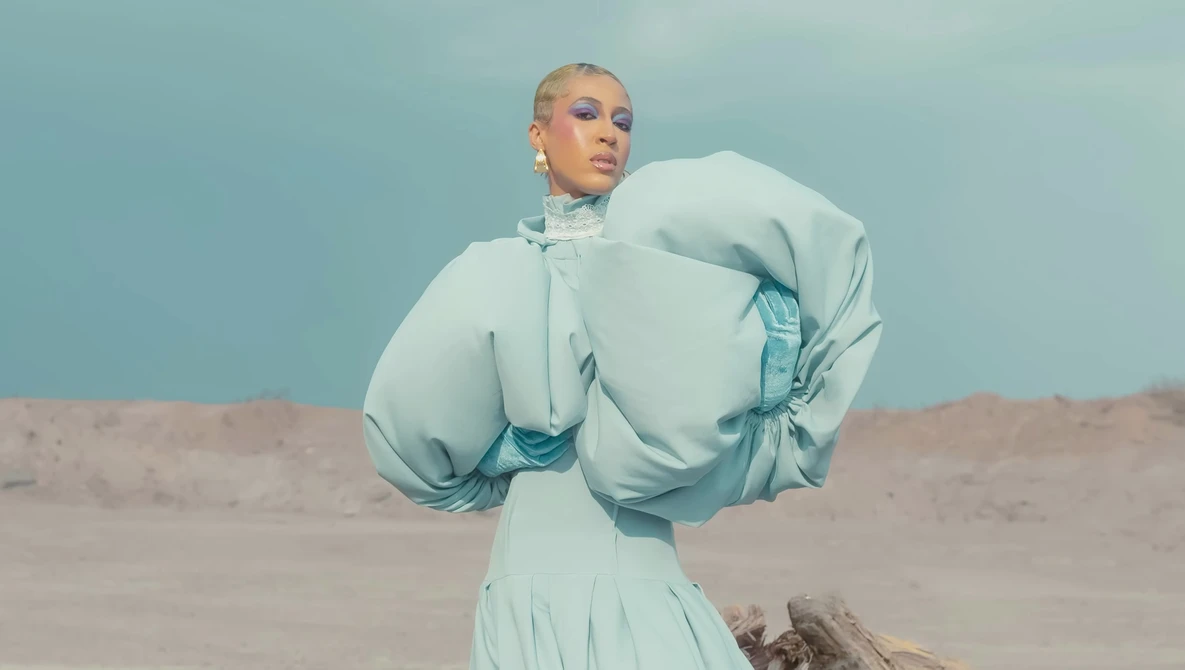 Person in flowing pale blue outfit with dramatic puffed sleeves standing in desert landscape against clear sky.