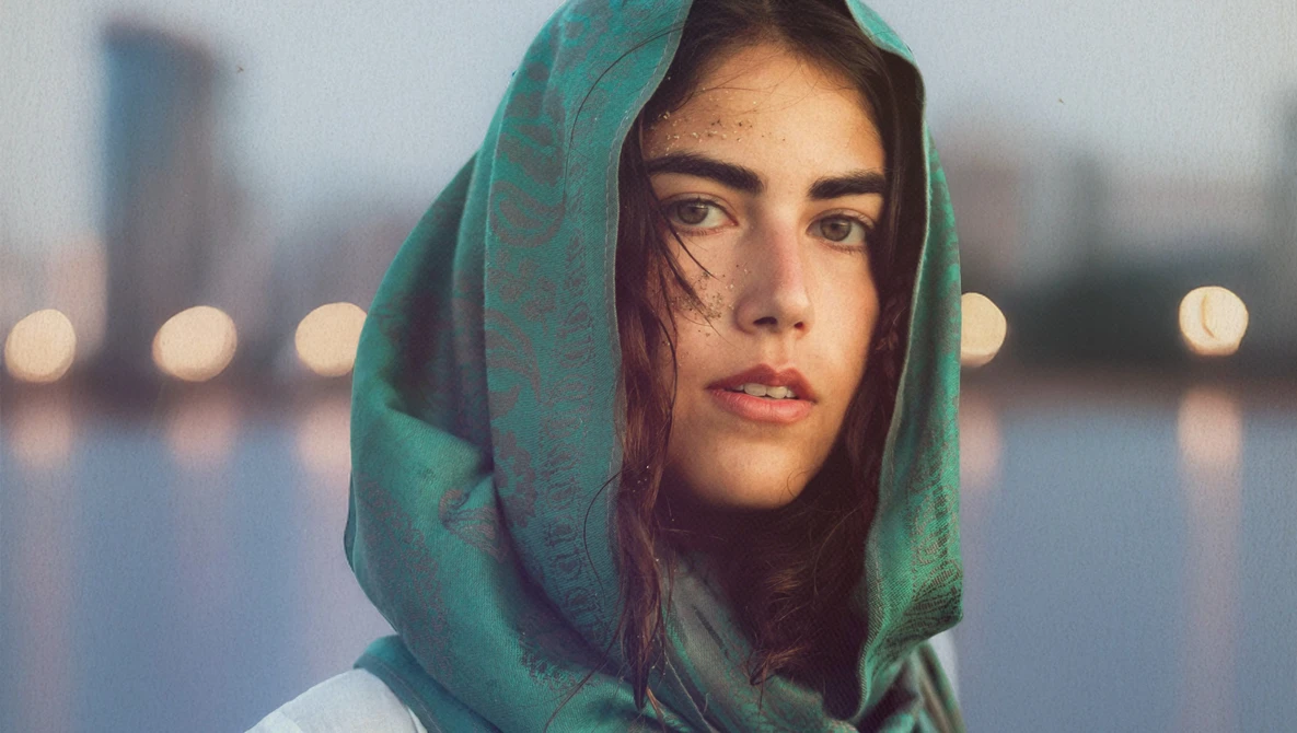 Woman wearing green headscarf looking at camera with urban waterfront and bokeh lights in background.