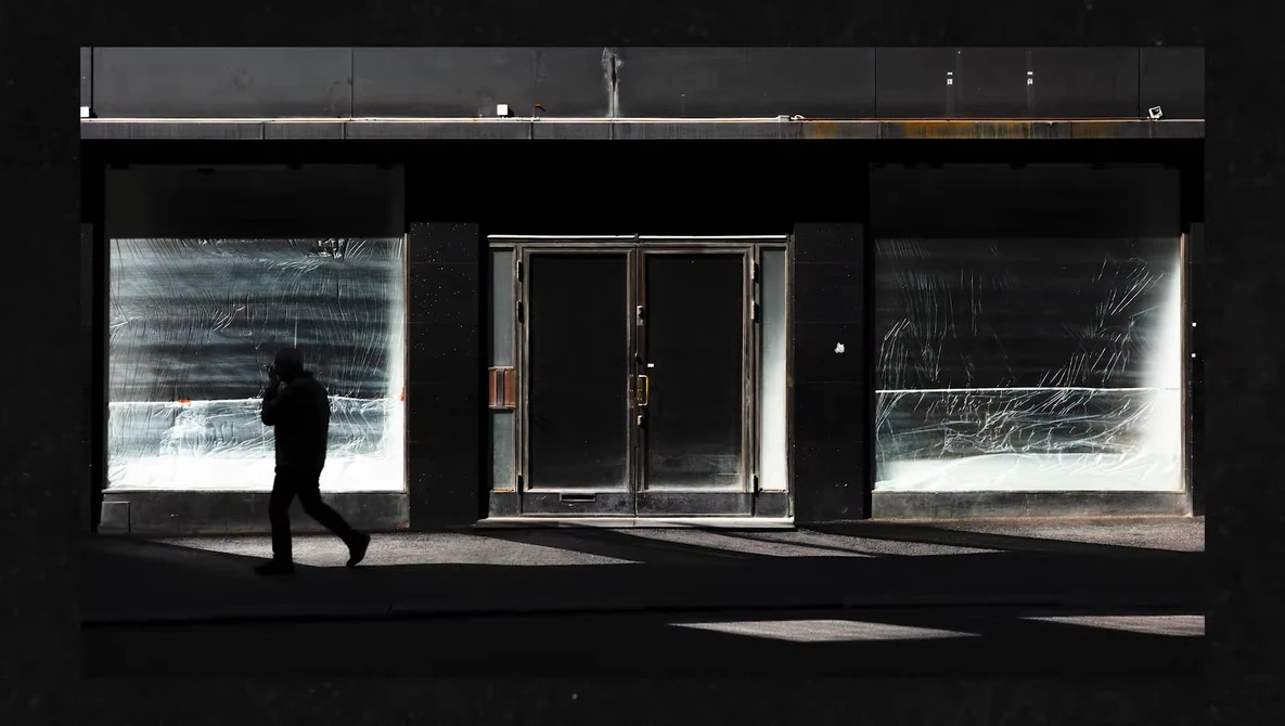 Silhouetted figure walking past storefront with illuminated window displays against dark facade.
