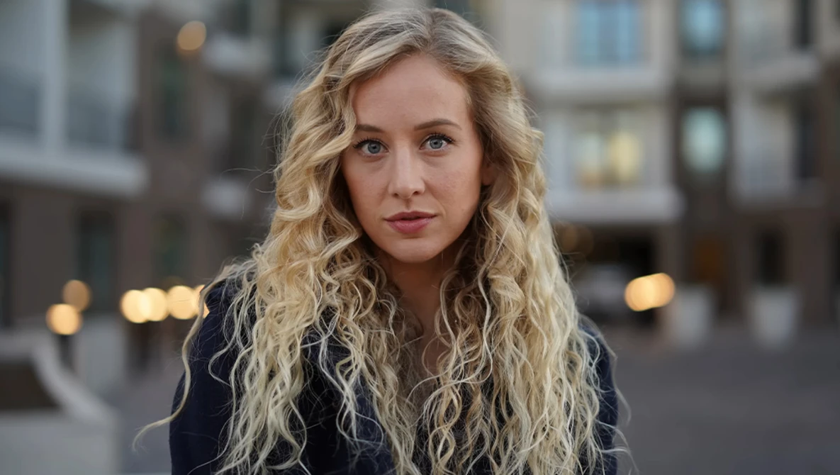 Woman with long blonde wavy hair looking at camera outdoors in urban setting.