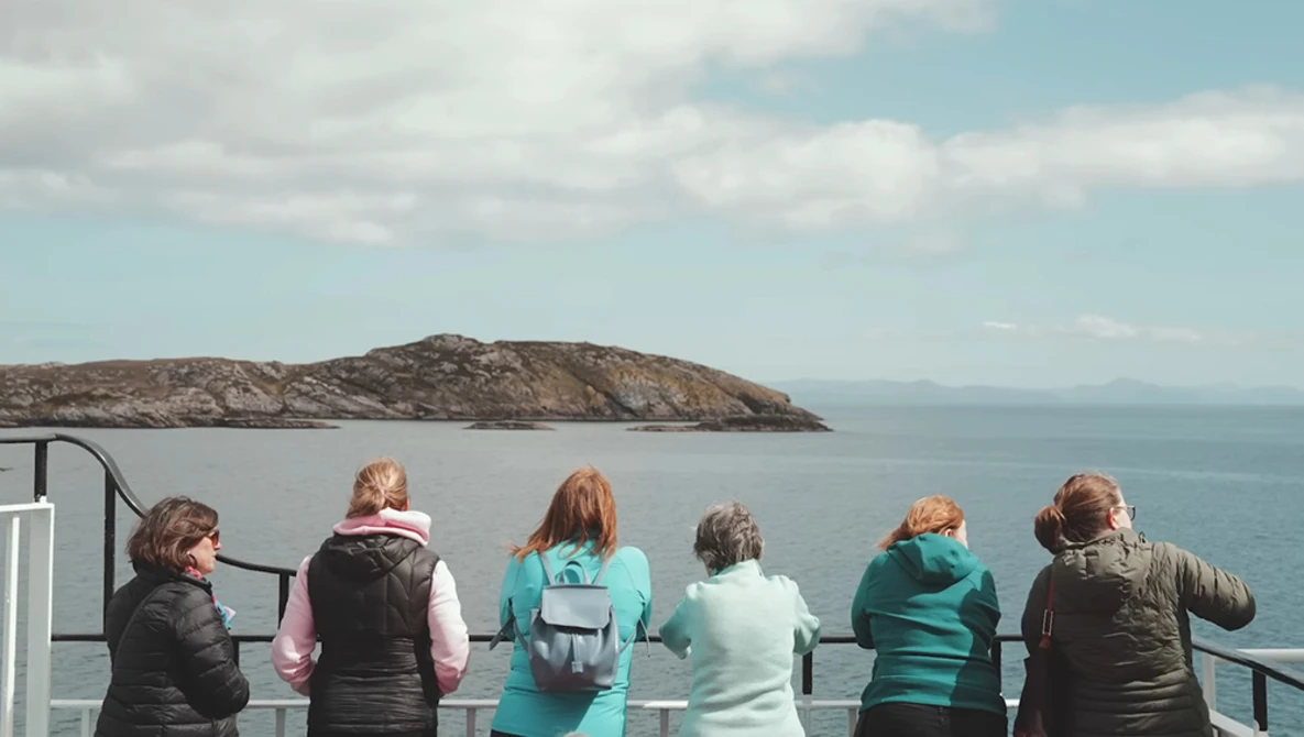 Six people viewing a rocky island from a boat railing on calm blue water.