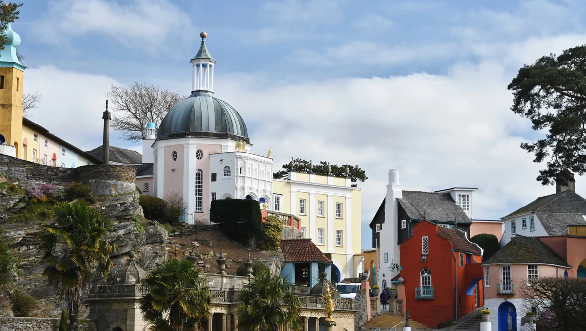 Hillside coastal town with white-domed chapel, yellow building, and colorful historic structures under partly cloudy sky.