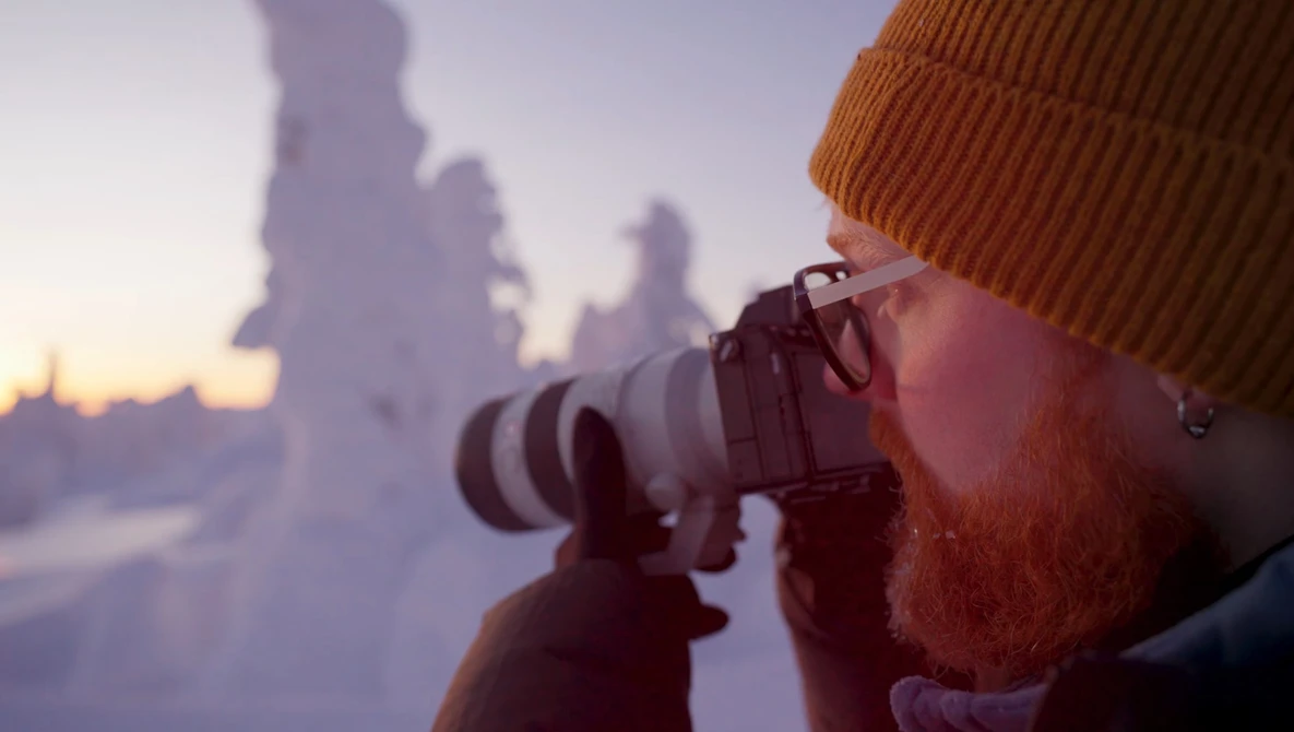 Bearded photographer with orange beanie holding telephoto lens to eye during golden hour, snow-covered landscape blurred in background.