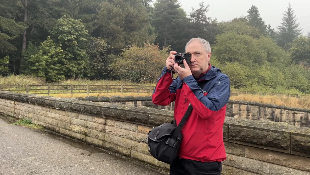 Male photographer in red and blue jacket holding a camera to his face while standing on a wooden boardwalk.