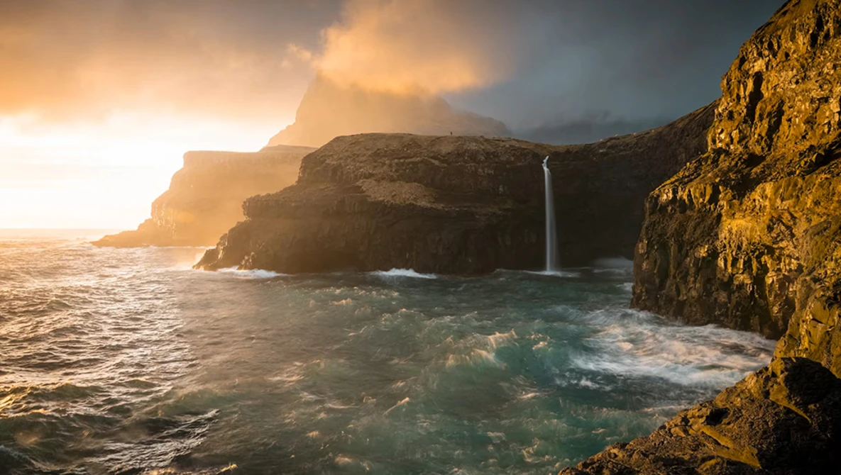 Waterfall cascading down golden cliffs into turquoise ocean waters during golden hour sunset.