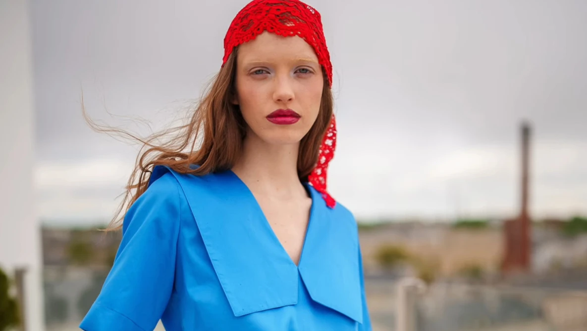 Woman wearing bright blue wrap blouse and red lace headwrap looking over shoulder outdoors.