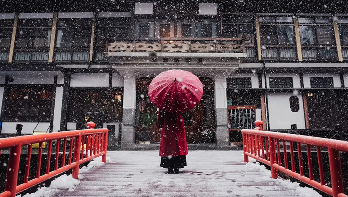 Person holding red umbrella walking across red bridge in heavy snowfall toward traditional architecture.