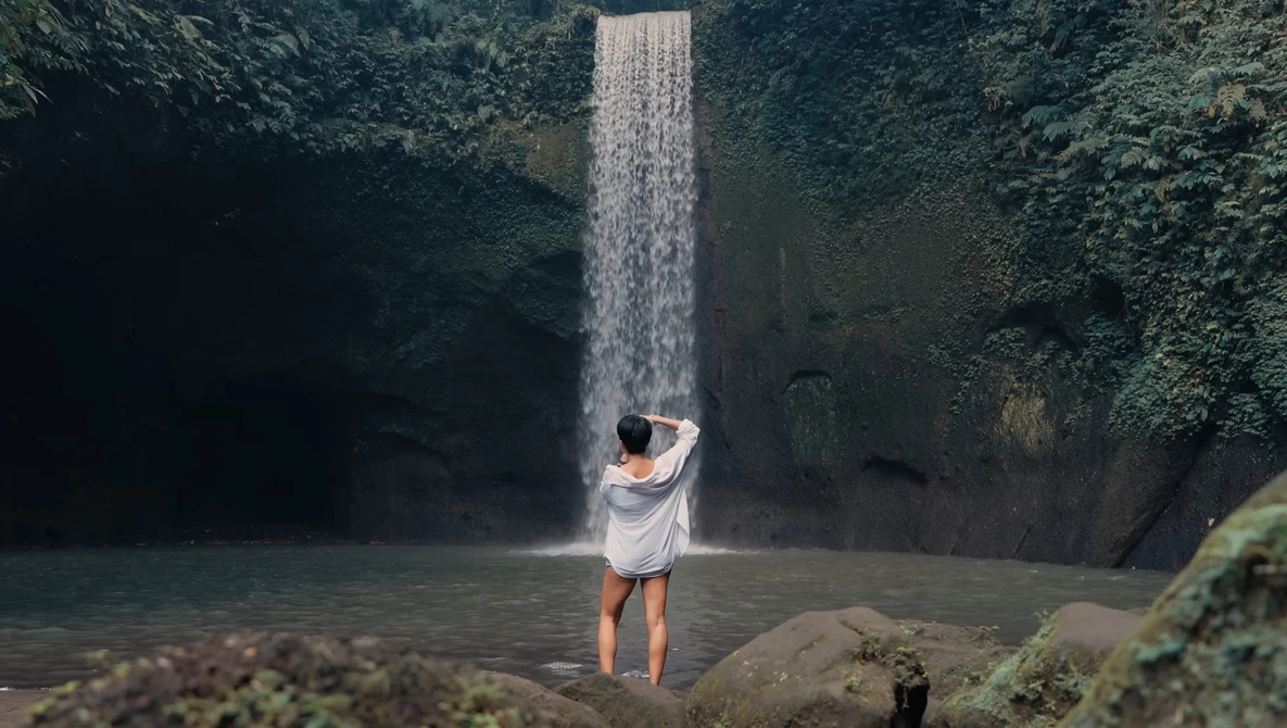 Person in white shirt standing in shallow water beneath a tall waterfall surrounded by lush vegetation.