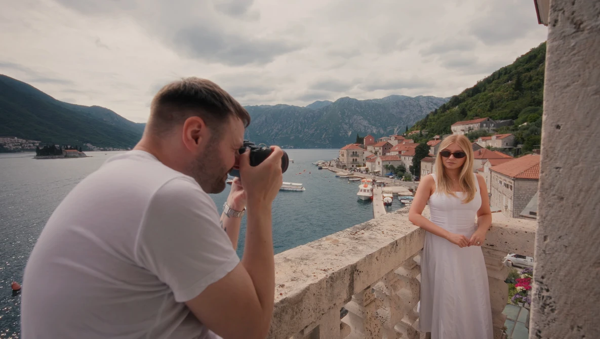 Photographer shoots portrait of woman in white dress against coastal bay backdrop.