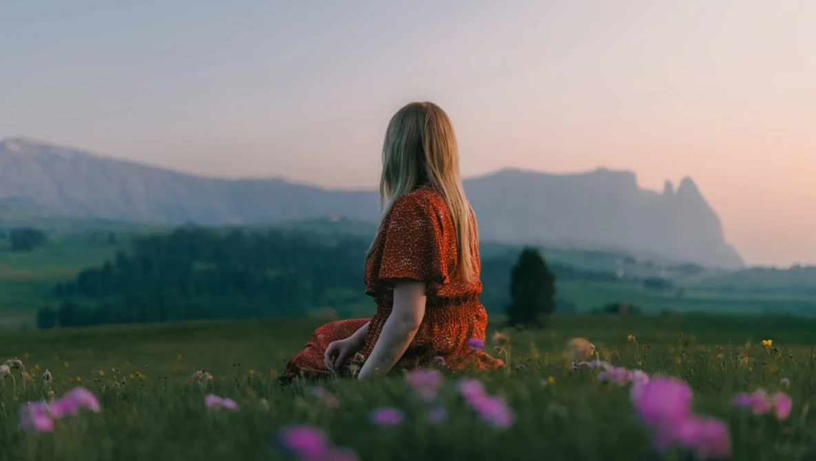 Woman in rust-colored dress sitting in wildflower meadow facing distant mountains at sunset.