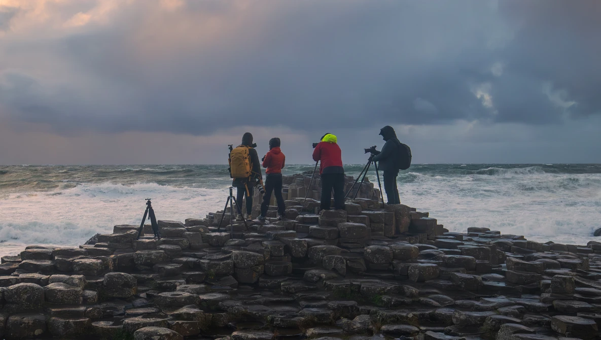 Four people with cameras and tripods standing on rocky coastal outcrop during overcast conditions.