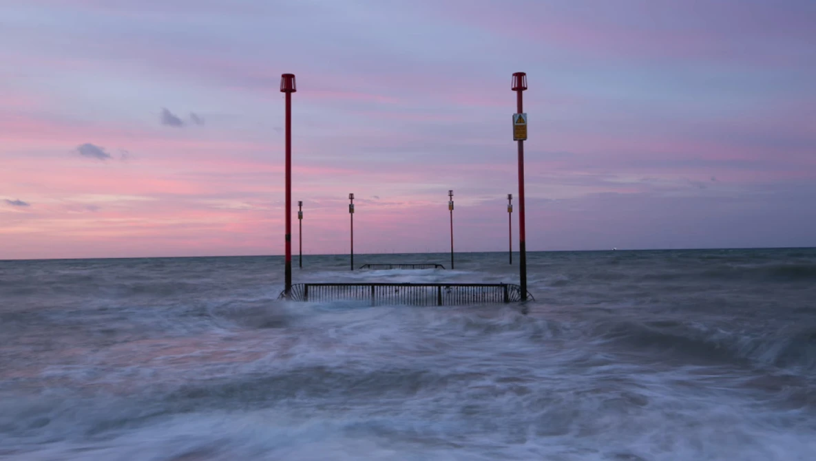 Wooden pier structure with tall posts standing in shallow ocean waves at dusk, with pink and purple sky.