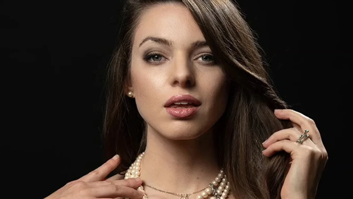 Woman with long dark hair wearing pearl necklace and rings against black background.
